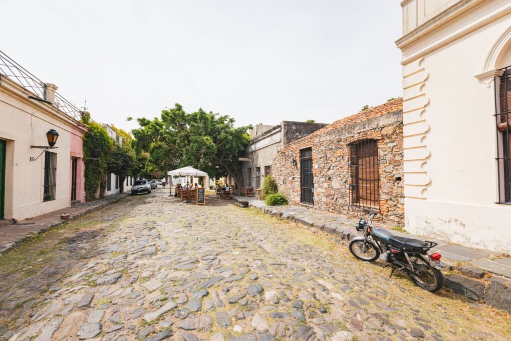 Rua pavimentada com pedras em uma cidade histórica com prédios em tons pastel, árvores e um café ao ar livre sob um guarda-sol branco Avenida General Flores em Colônia do Sacramento
