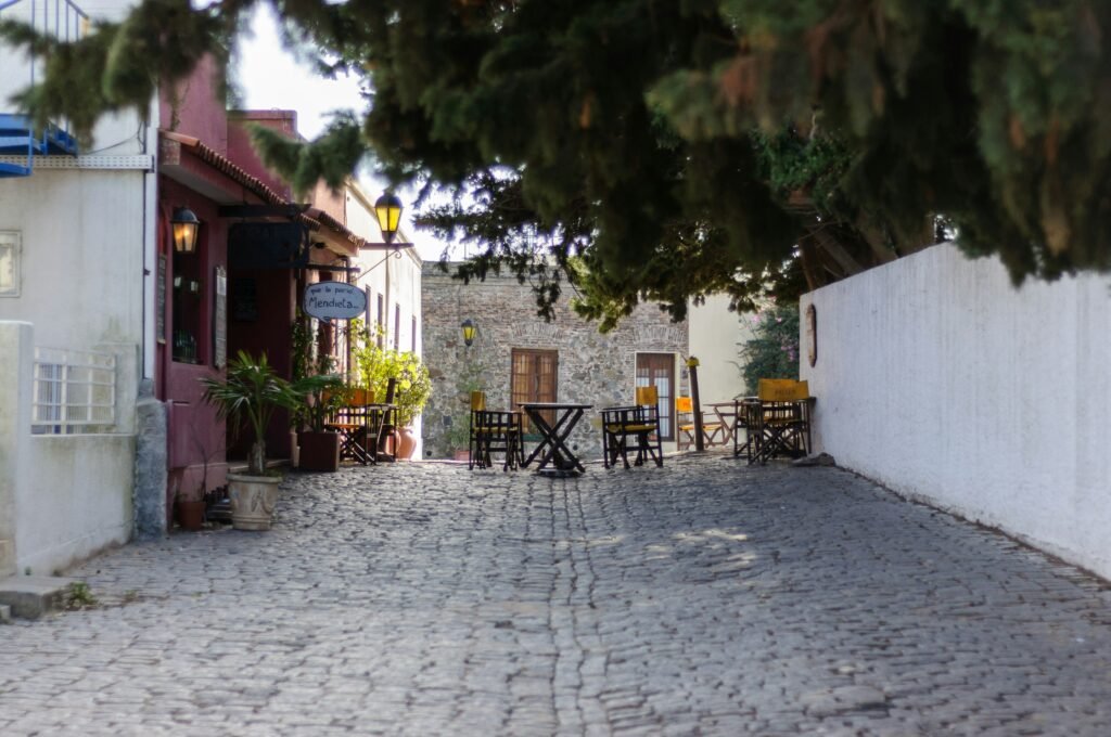 Rua de paralelepípedos em um beco ensolarado com mesas e cadeiras de café ao ar livre, ladeada por fachadas coloridas e uma parede branca Avenida General Flores em Colônia do Sacramento