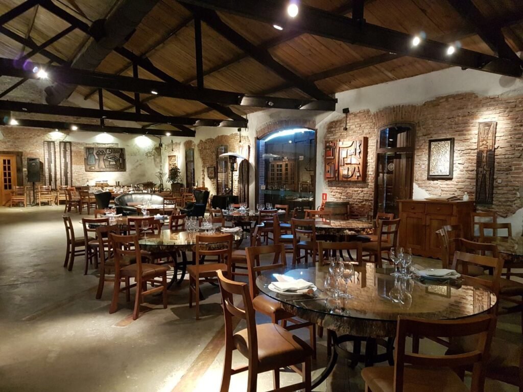 Restaurante rústico e aconchegante com paredes de tijolos, vigas de madeira expostas e mesas redondas preparadas para jantar com copos.
