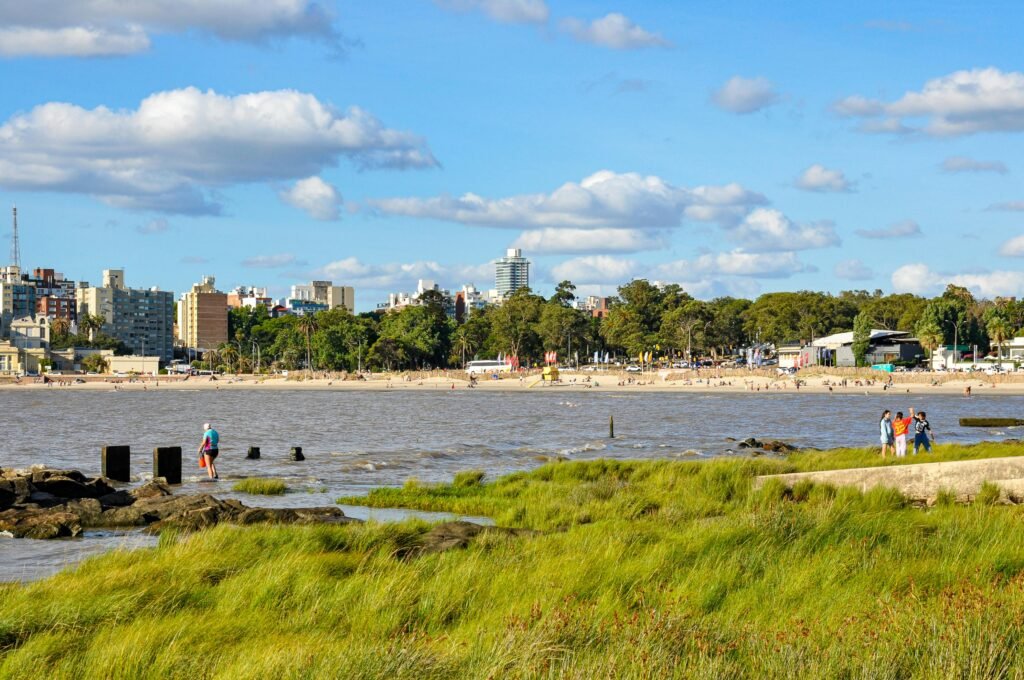 Vista da cidade ao fundo, com uma praia de areia, dunas gramadas e pessoas passeando à beira-mar em um dia ensolarado Farol de Punta Carretas em Montevideo