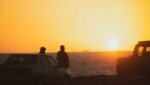 Silhuetas de duas pessoas em pé junto a carros estacionados na margem de um rio ao pôr do sol, com um céu laranja brilhante e água calma ao fundo Farol de Punta Carretas em Montevideo