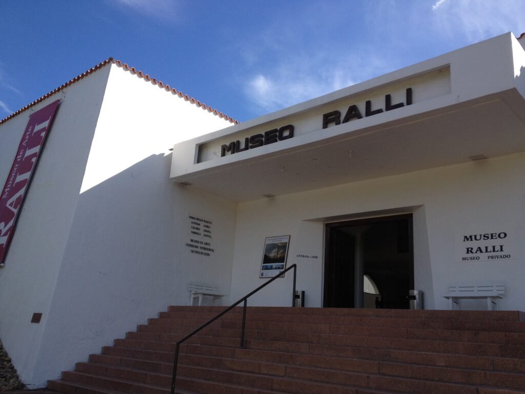 Entrada do museu com prédio branco, degraus marrons e placa 'MUSEO RALLI' sob um céu azul.