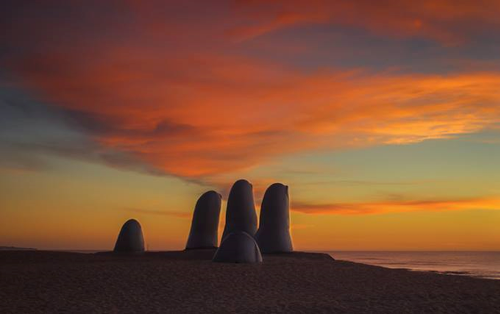 Silhuetas de pilares rochosos em forma de dedos numa praia ao pôr do sol, com céu alaranjado e águas calmas.