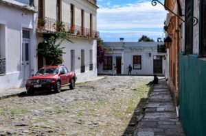 Uma rua de paralelepípedos em Colonia del Sacramento com grama entre as pedras em uma cidade histórica. À esquerda, uma caminhonete vermelha está estacionada em frente a casas brancas de estilo colonial com varandas de ferro. Ao fundo, pedestres caminham pela rua estreita sob um céu azul com nuvens leves. À direita, uma parede verde vibrante e uma luminária de metal trazem cor à cena.