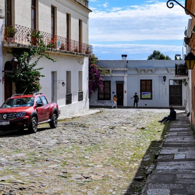 Uma rua de paralelepípedos em Colonia del Sacramento com grama entre as pedras em uma cidade histórica. À esquerda, uma caminhonete vermelha está estacionada em frente a casas brancas de estilo colonial com varandas de ferro. Ao fundo, pedestres caminham pela rua estreita sob um céu azul com nuvens leves. À direita, uma parede verde vibrante e uma luminária de metal trazem cor à cena.