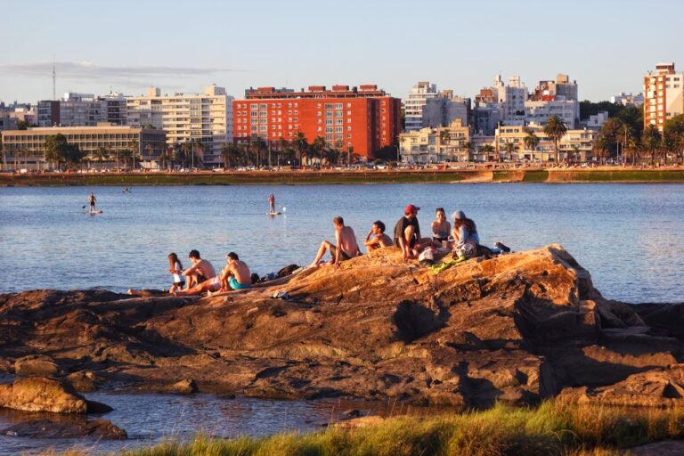 Pessoas sentadas em rochas à beira-mar em Montevidéu sob a luz do fim de tarde. Água calma com pessoas em pranchas de stand-up paddle e prédios da cidade ao fundo sob um céu limpo.