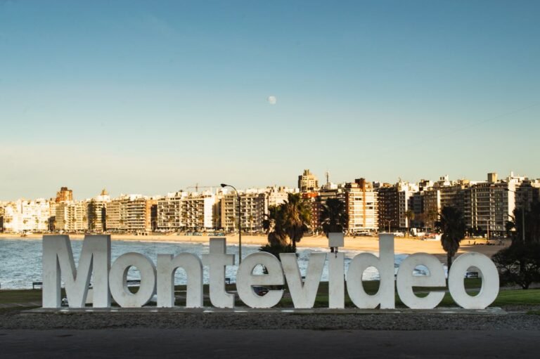Letreiro icônico de "Montevideo" em grandes letras brancas, localizado na Rambla de Pocitos, Uruguai. Ao fundo, a orla da Playa de los Pocitos com prédios residenciais, palmeiras e o mar sob um céu azul claro com a lua visível.