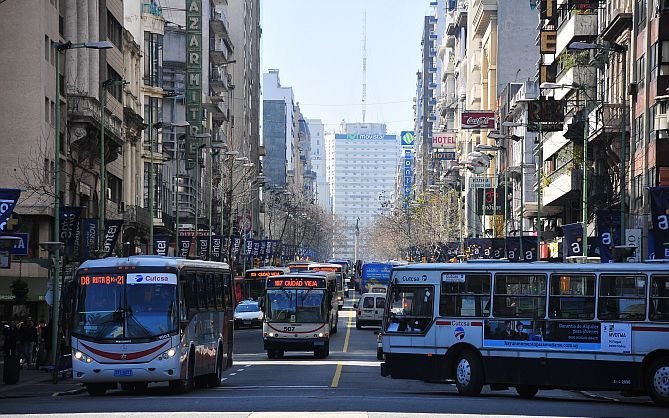 Vista de rua urbana movimentada em Montevidéu sob luz solar. Vários ônibus de grande porte brancos e cinzas ocupam as faixas centrais de uma avenida larga, ladeada por edifícios altos com letreiros de hotéis, Coca-Cola e publicidade da Movistar ao fundo.