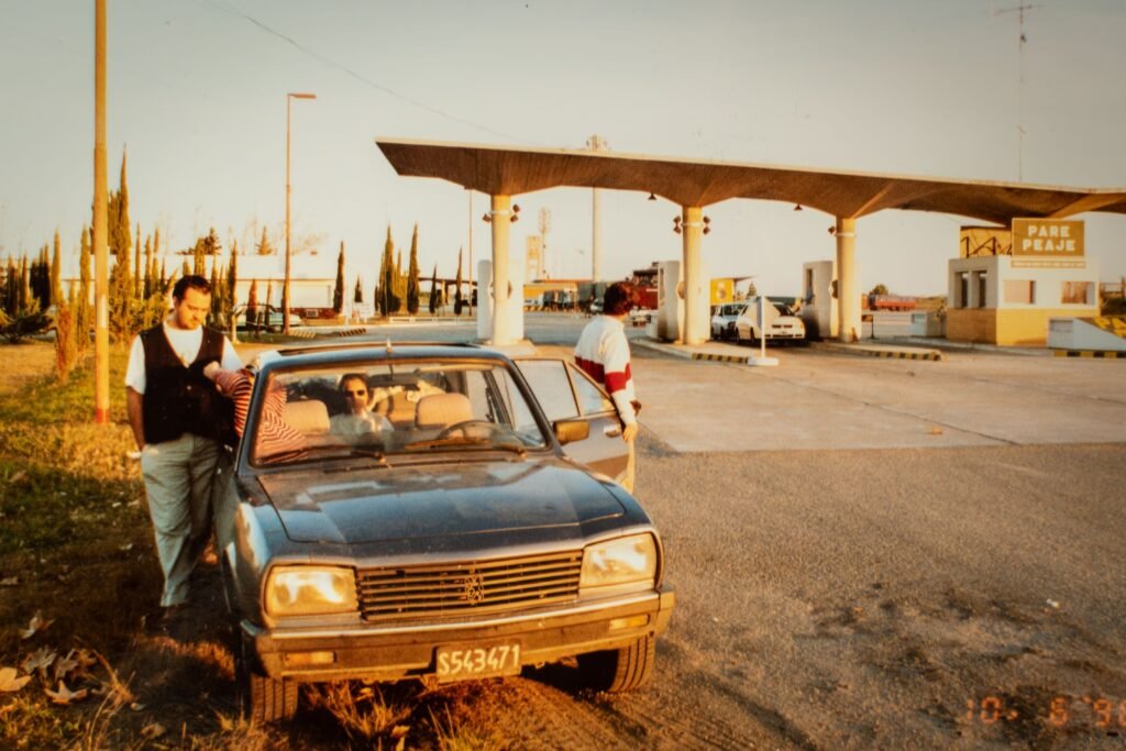 Um homem de colete está ao lado de um conversível azul em um posto de gasolina antigo, outra pessoa perto do carro, com uma grande cobertura e bombas de combustível ao fundo, sob a luz do pôr do sol Noite da Nostalgia no Uruguai