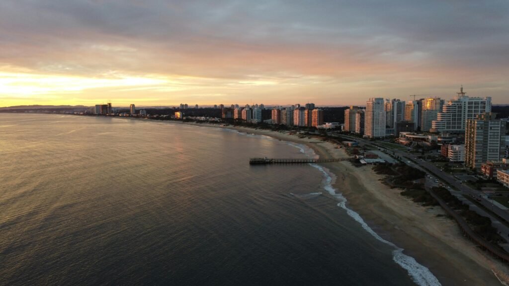 Vista da orla de uma cidade ao pôr do sol, com uma fileira de prédios altos ao longo da praia e um píer que se estende mar adentro Noite da Nostalgia no Uruguai