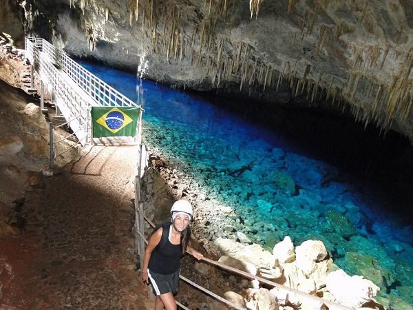Adriana sorridente usando capacete de proteção caminha por uma passarela dentro de uma caverna iluminada. Ao lado da trilha, há um lago de águas extremamente azuis e cristalinas, onde se veem rochas no fundo. O teto da caverna está repleto de estalactites. Uma bandeira do Brasil está pendurada no corrimão da passarela metálica ao fundo.