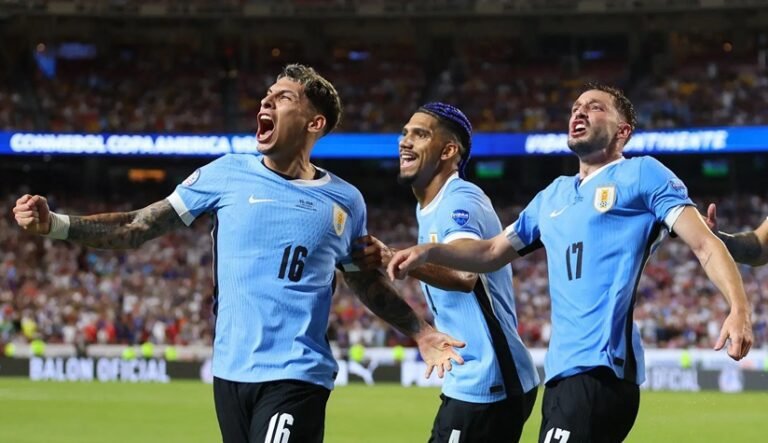Jogadores da seleção uruguaia, incluindo Mathias Olivera (16) e Matías Viña (17), celebram um gol com euforia durante uma partida da CONMEBOL Copa América. A imagem mostra os atletas vestindo o uniforme oficial azul celeste em um estádio lotado, simbolizando a paixão e a vibração do futebol no Uruguai atual.