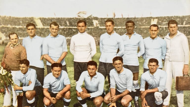 Fotografia colorizada de 1928 mostrando o time de futebol do Uruguai abraçado e ajoelhado no gramado, celebrando o bicampeonato olímpico em um estádio cheio.
