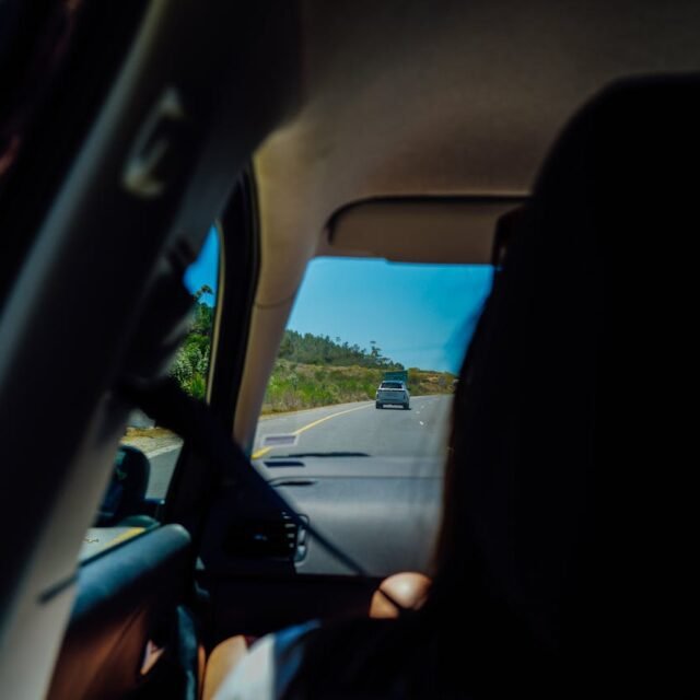 Fotografia tirada do banco traseiro de um carro, mostrando a perspectiva de um passageiro durante uma viagem. Através do para-brisa, vê-se uma estrada asfaltada que serpenteia por colinas verdes sob um céu azul limpo. Um veículo cinza está à frente na estrada. O interior do carro está em sombras, com parte da silhueta de um passageiro e o cinto de segurança visíveis em primeiro plano.