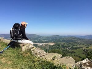 Adriana de costas, sentada em uma pedra no alto de uma montanha, com mochila de trilha e bastão de caminhada, durante viagem solo pelo Uruguai. Ela olha para uma paisagem ampla de vales verdes, colinas e céu azul limpo.