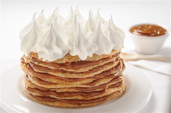 Stack of caramel-drizzled pancakes with generous whipped cream on top, served on a white plate; caramel sauce in a small cup in the background.