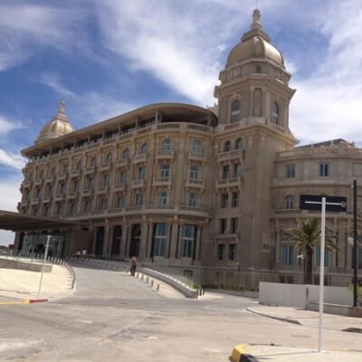 Beige seaside hotel or landmark building with domed towers and arched windows under a blue sky.