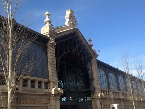 Historic stone train-station style building with an ornate arched entrance, decorative pinnacles, and large glass windows against a blue sky.