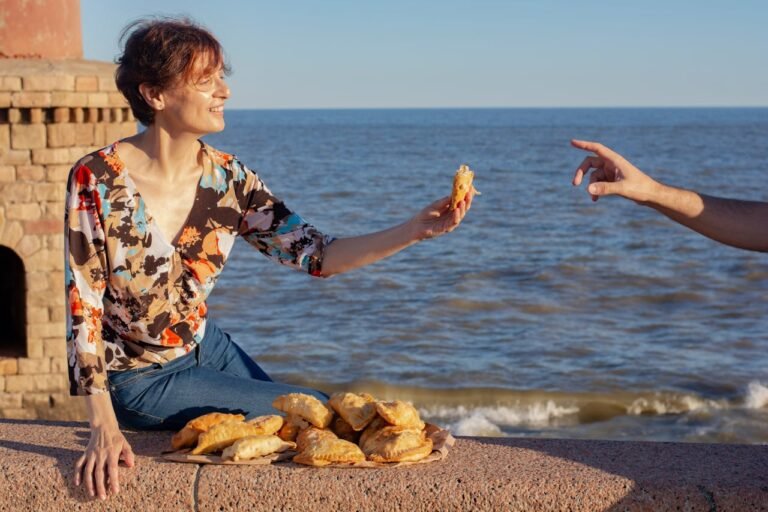 Fotografia diurna de uma mulher com blusa floral sentada em um parapeito de pedra à beira-mar. Ela oferece uma empanada para uma mão que aparece lateralmente. No muro, há diversas empanadas empilhadas sobre um papel pardo, com o horizonte do Rio da Prata ao fundo sob céu limpo