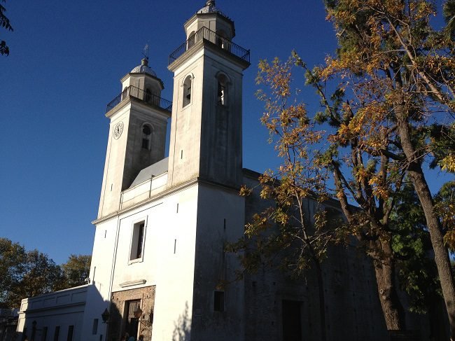 Fotografia diurna de uma igreja histórica branca com duas torres sineiras altas sob céu azul limpo. Uma das torres possui um relógio. Árvores com folhas secas emolduram a lateral da construção em Colonia del Sacramento.