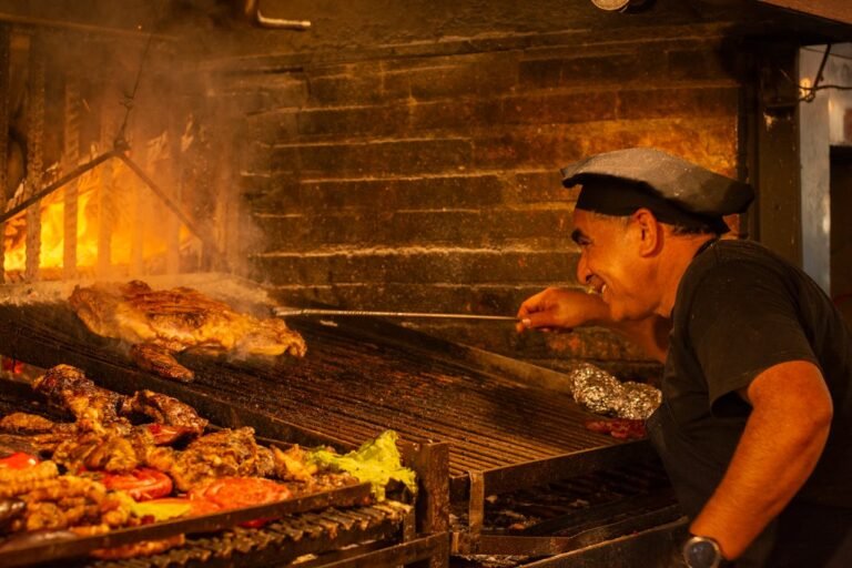 Um parrilheiro sorridente manuseia cortes de carne em uma autêntica parrilla uruguaia com fundo de tijolos e braseiro ardente. A grelha está repleta de comidas típicas do Uruguai sendo preparadas de forma tradicional, uma das principais atrações para quem busca o que fazer no Uruguai.