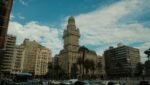 Vista panorâmica da Plaza Independencia em Montevidéu, Uruguai, com o imponente edifício Palácio Salvo em destaque central. A imagem captura a arquitetura clássica e eclética do marco histórico, cercado por palmeiras, edifícios modernos, trânsito urbano e um ônibus do sistema local sob um céu azul com nuvens esparsas.