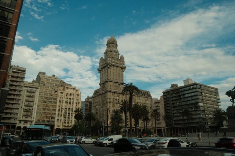 Vista panorâmica da Plaza Independencia em Montevidéu, Uruguai, com o imponente edifício Palácio Salvo em destaque central. A imagem captura a arquitetura clássica e eclética do marco histórico, cercado por palmeiras, edifícios modernos, trânsito urbano e um ônibus do sistema local sob um céu azul com nuvens esparsas.