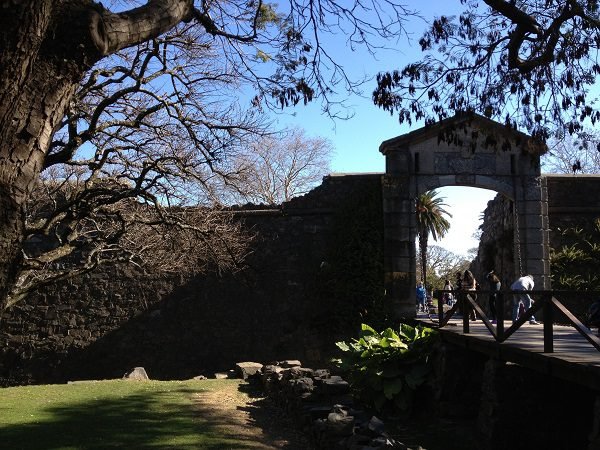 Vista da histórica Puerta de la Ciudadela (Portón de Campo) em Colonia del Sacramento, Uruguai. A imagem destaca o portão de pedra com arco, a ponte de madeira e as muralhas antigas que preservam o charme colonial do centro histórico, um dos destinos imperdíveis para turistas brasileiros.