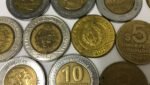 Várias moedas uruguaias de 5 e 10 pesos espalhadas em uma superfície clara. As moedas bimetálicas de 10 pesos estão em destaque ao lado das moedas douradas de 5 pesos.