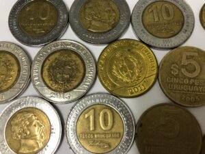 Várias moedas uruguaias de 5 e 10 pesos espalhadas em uma superfície clara. As moedas bimetálicas de 10 pesos estão em destaque ao lado das moedas douradas de 5 pesos.