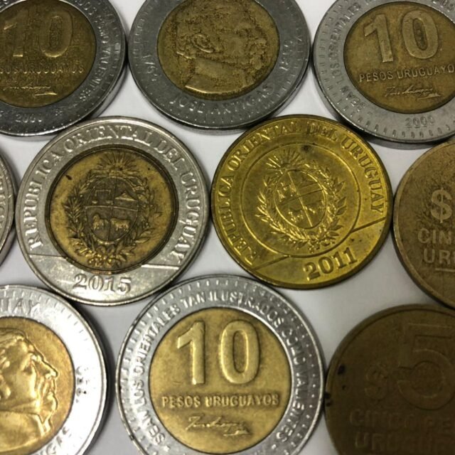 Várias moedas uruguaias de 5 e 10 pesos espalhadas em uma superfície clara. As moedas bimetálicas de 10 pesos estão em destaque ao lado das moedas douradas de 5 pesos.