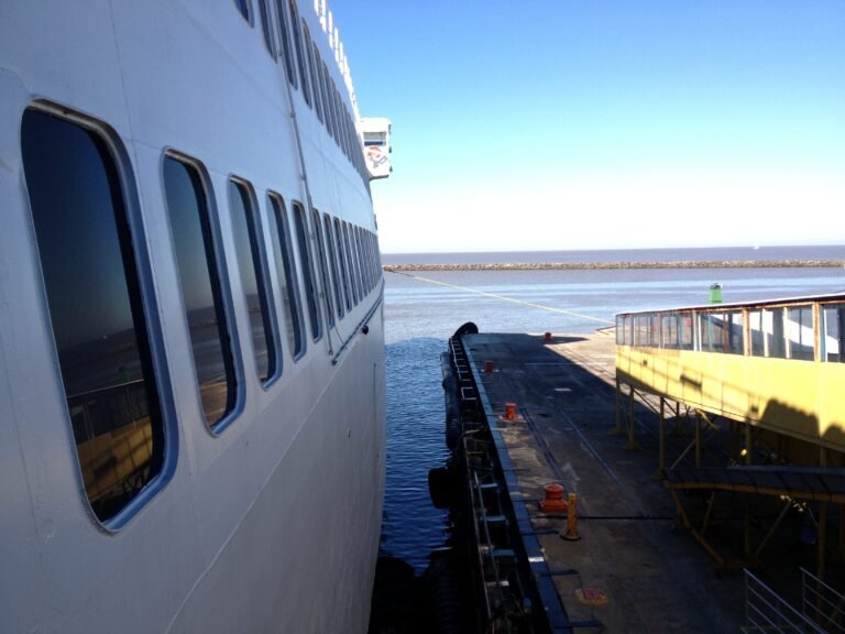 Vista detalhada da lateral de uma balsa rápida (ferry) da Buquebus, mostrando as linhas de janelas ovais e o casco branco imponente atracado ao dock. No terminal, vê-se a passarela amarela de embarque, o quebra-mar de pedras e as águas do Río de la Plata sob o céu azul, marcando o início da travessia.