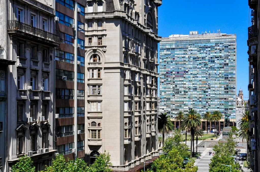 Fileira de edifícios ornamentados de pedra alinhados em uma rua da cidade, com palmeiras e uma torre de vidro moderna ao fundo, sob um céu azul. Banco de la República Oriental del Uruguay