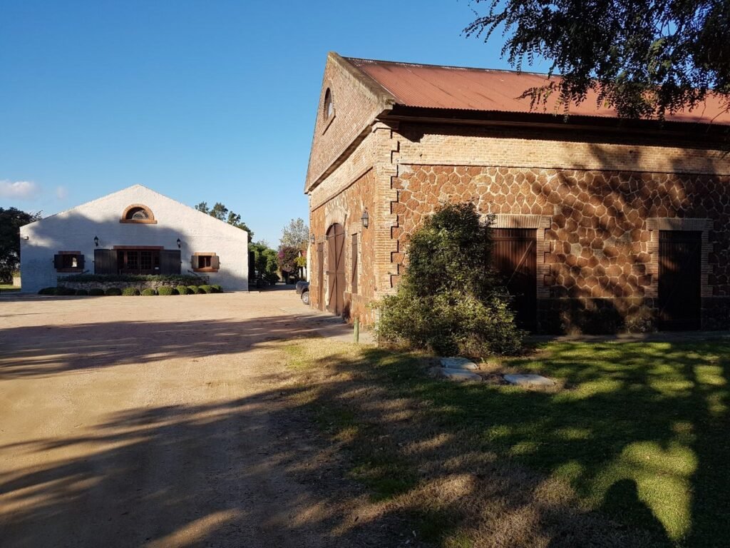 Two rustic farm buildings under a clear blue sky: a white stucco house with arched window and shutters on the left, and a brown stone barn with a red roof on the right.