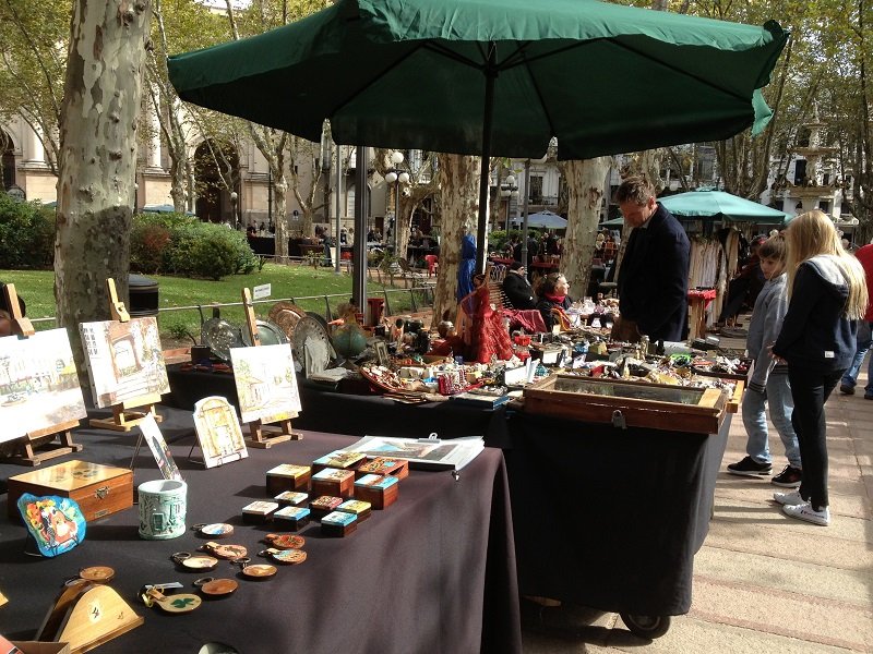 Outdoor market scene with vendors under a large green umbrella selling handmade crafts and trinkets on tables while shoppers browse.