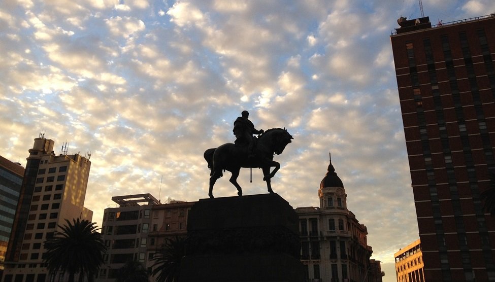 Equestrian statue silhouetted against a cloudy sunset with surrounding city buildings in a plaza behind it.