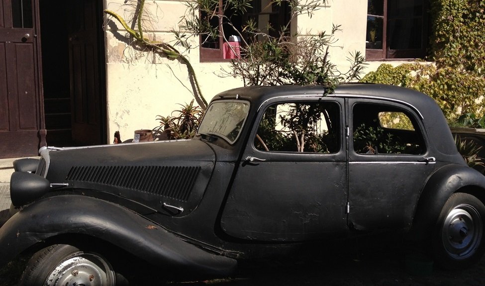 Classic black vintage coupe with rounded fenders parked beside a beige building with greenery in the background