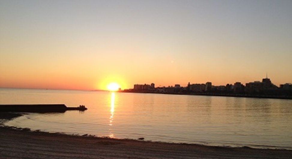 Sunset over a calm sea with a distant city skyline and a small jetty extending into the water.