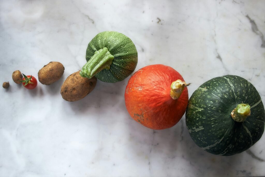 Assorted vegetables on a marble surface: green squash, orange pumpkin, striped green pumpkin, small potatoes, and a cherry tomato.