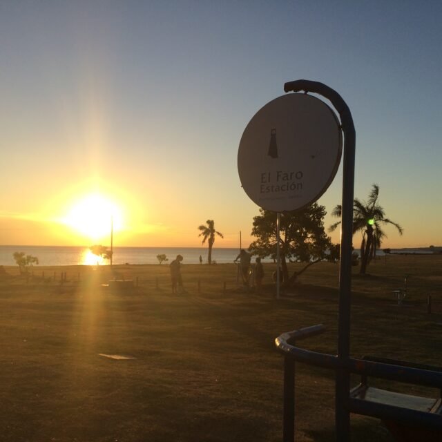 Pôr do sol sobre a praia com uma placa circular com os dizeres 'El Faro Estación' e silhuetas de pessoas e palmeiras ao perto, brilho quente na água melhores hotéis em Montevideo