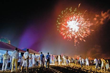 People gather on a sandy beach watching a bright fireworks display over a dark sky at night.