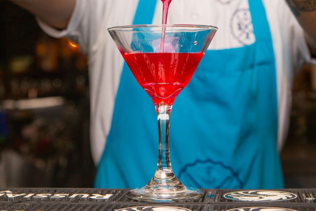 Um coquetel vermelho sendo servido em uma taça de martini, posicionada sobre um balcão de bar, com um barman de avental azul ao fundo. Burlesque Bar em Montevideo