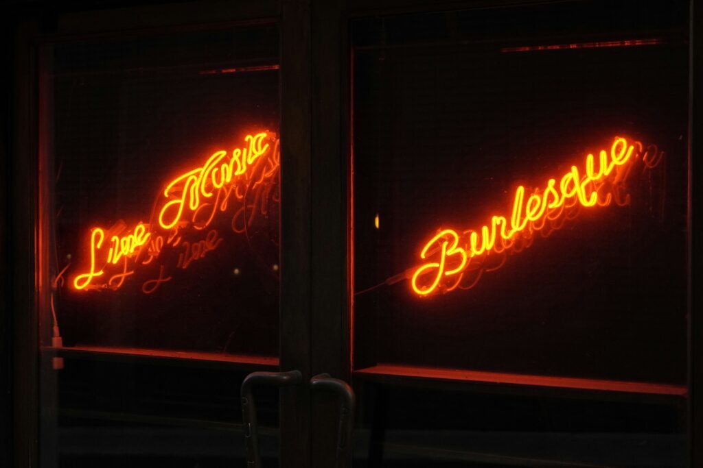 Um letreiro de néon laranja brilhante com a palavra "Bebidas" reluzindo na vitrine escura de uma loja, com reflexos nas portas de vidro à noite. Burlesque Bar em Montevideo