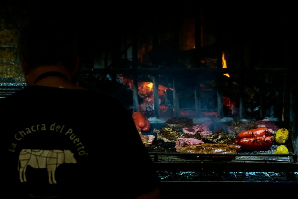 Costas de uma pessoa vestindo uma camisa escura, em pé em frente a uma churrasqueira com diversos tipos de carne assando sobre chamas intensas Cabildo de Montevideo