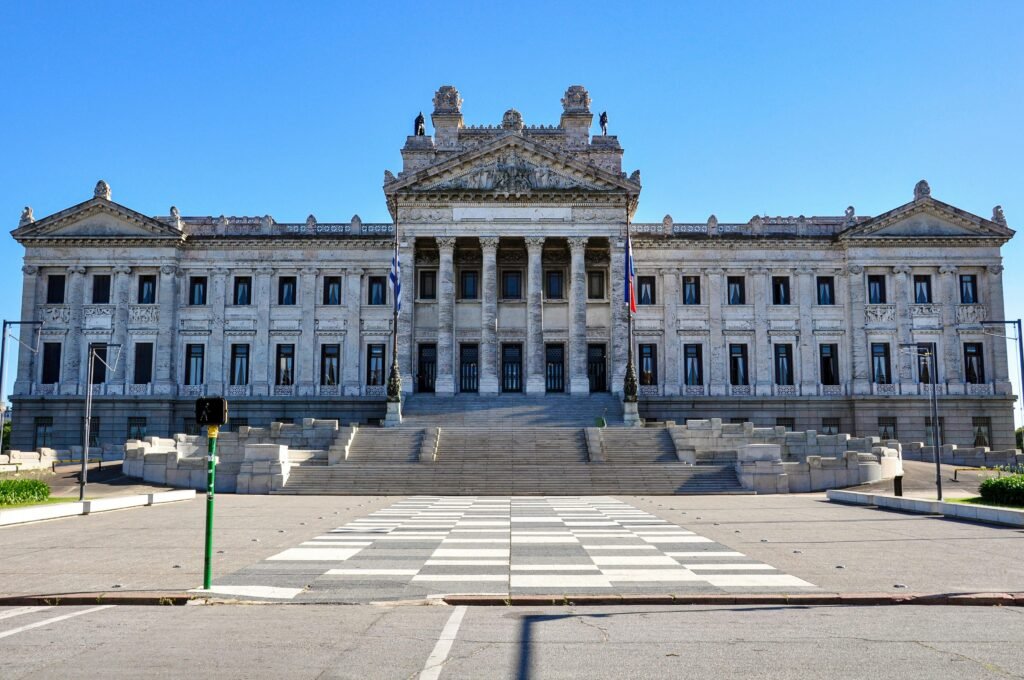Grande edifício governamental neoclássico com altas colunas, frontão ornamentado, estátuas no topo, bandeiras e ampla escadaria sob um céu azul Cabildo de Montevideo
