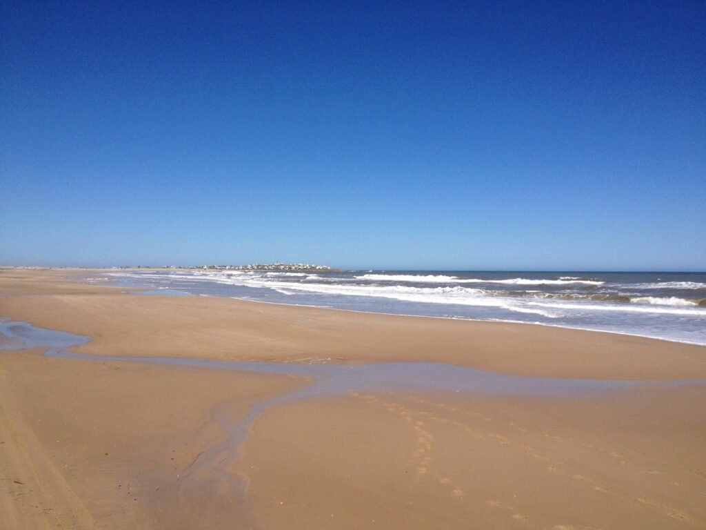 Praia de areia extensa, com oceano azul e calmo e uma cidade distante no horizonte, sob um céu azul e límpido.