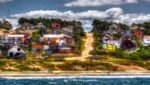 Vila costeira com casas coloridas situadas em uma colina, com vista para uma praia de areia e o oceano azul.