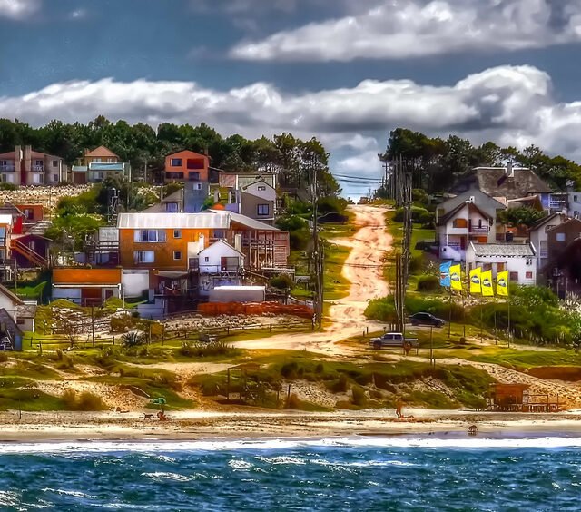 Vila costeira com casas coloridas situadas em uma colina, com vista para uma praia de areia e o oceano azul.