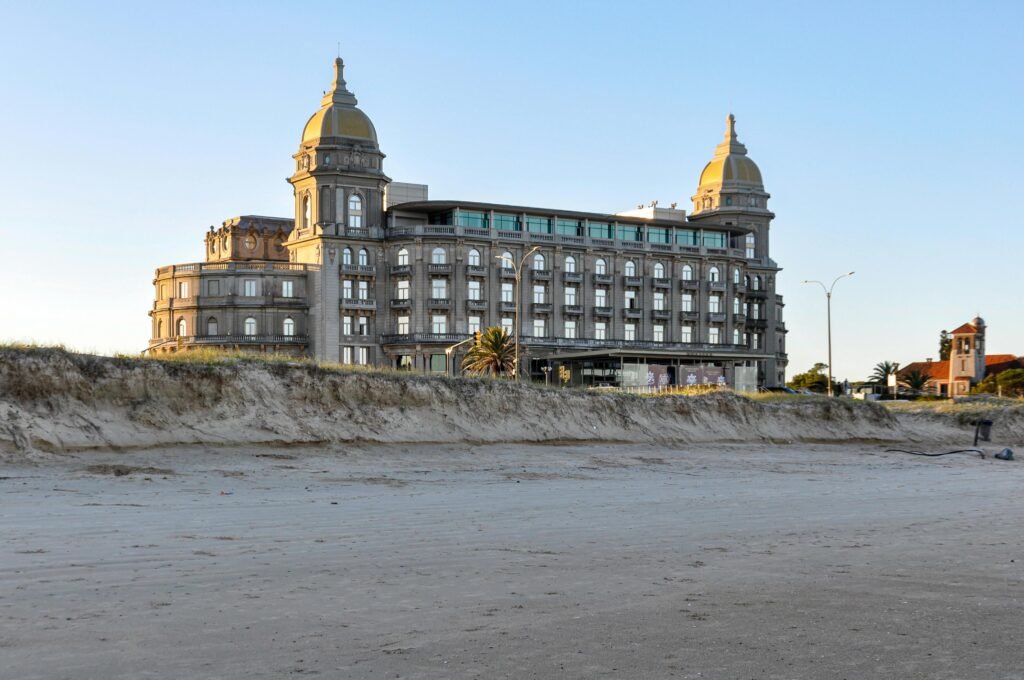 A imagem de um edifício histórico de pedra com duas torres em forma de cúpula, situado numa praia de areia e com um céu azul límpido ao pôr do sol, não está presente, mas o texto descreve bem a cena Cassino Carrasco em Montevideo