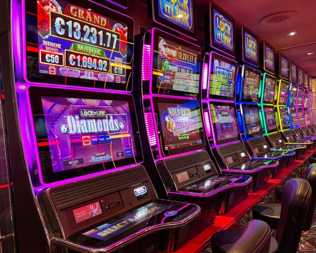 Fileira de máquinas caça-níqueis coloridas em um cassino, com telas brilhantes exibindo jackpots e detalhes em rosa neon/roxo Cassino Victoria Plaza em Montevideo